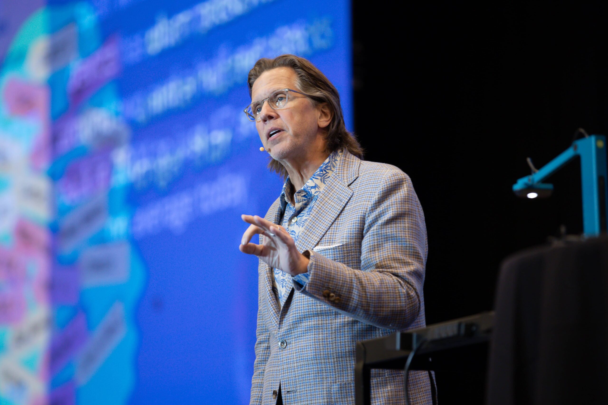 A man wearing glasses and a patterned blazer speaks on stage, gesturing with his hand while addressing an audience. He wears a headset microphone, and a large, colorful presentation screen is blurred in the background.
