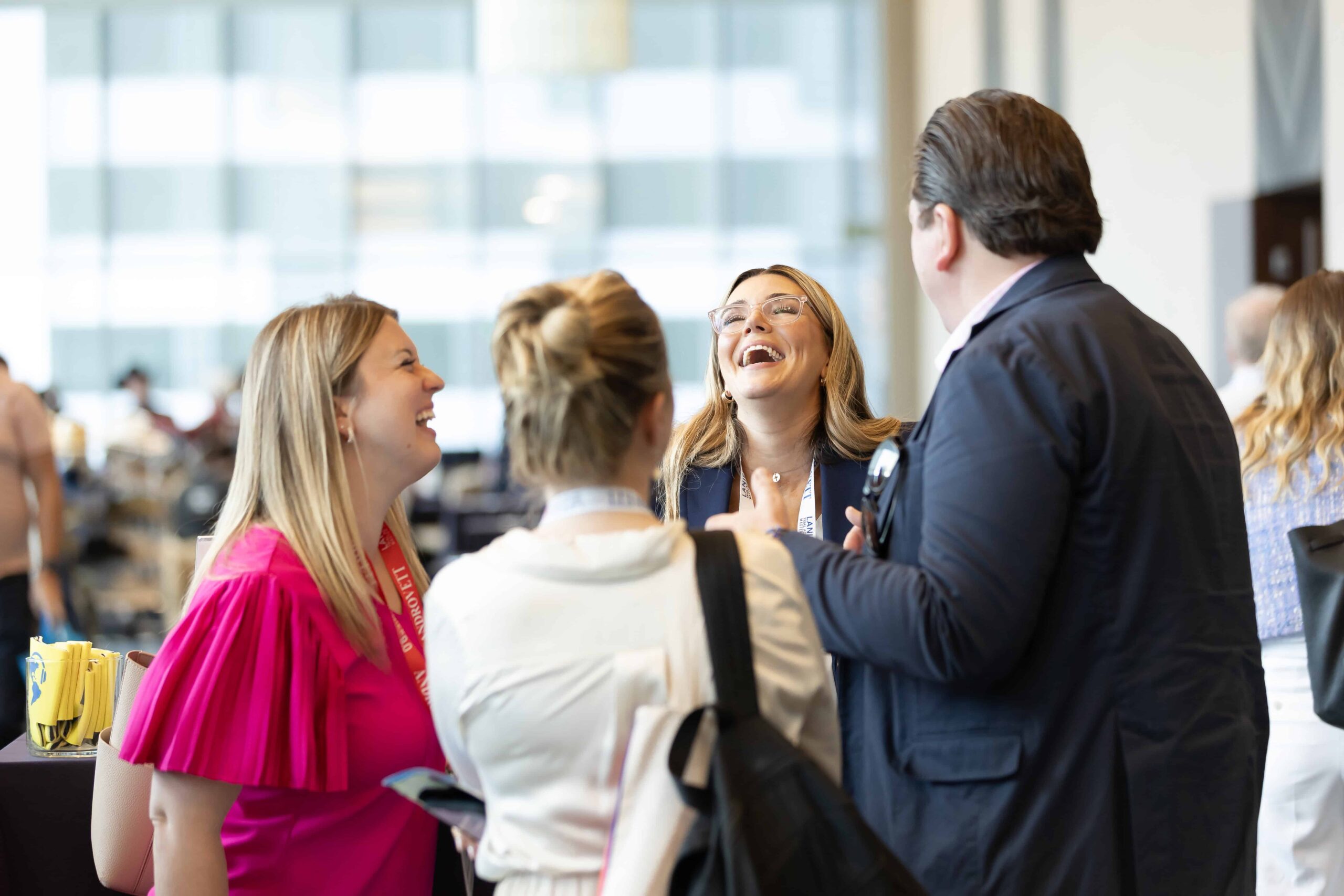 A group of four professionally dressed adults stand in a bright, modern indoor venue, laughing and chatting together during what appears to be a conference or networking event. One woman in a pink top faces the group, while another woman with glasses smiles broadly. A man in a suit stands with his back partially to the camera, and another woman with her hair in a bun carries a bag over her shoulder. The background shows a softly blurred crowd and large windows letting in natural light.