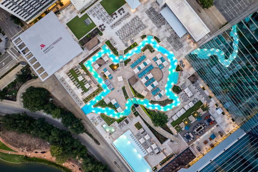 Aerial view of the Marriott Marquis Houston rooftop pool deck featuring its iconic Texas-shaped lazy river, lounge chairs, cabanas, and surrounding city buildings.